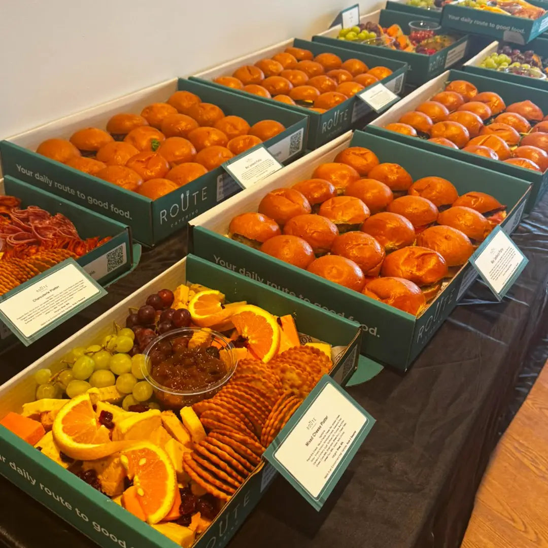 Multiple green ROUTE-branded gift boxes filled with assorted foods, including orange-colored round snacks, sliced oranges, grapes, crackers, and a small bowl of dried fruit, arranged on a black tablecloth.