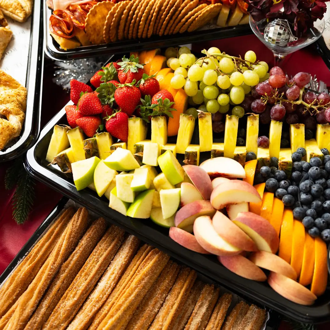 A black catering platter filled with assorted fruit slices, grapes, strawberries, blueberries, and pineapple sticks, alongside trays of churritos and crackers in a party setting.